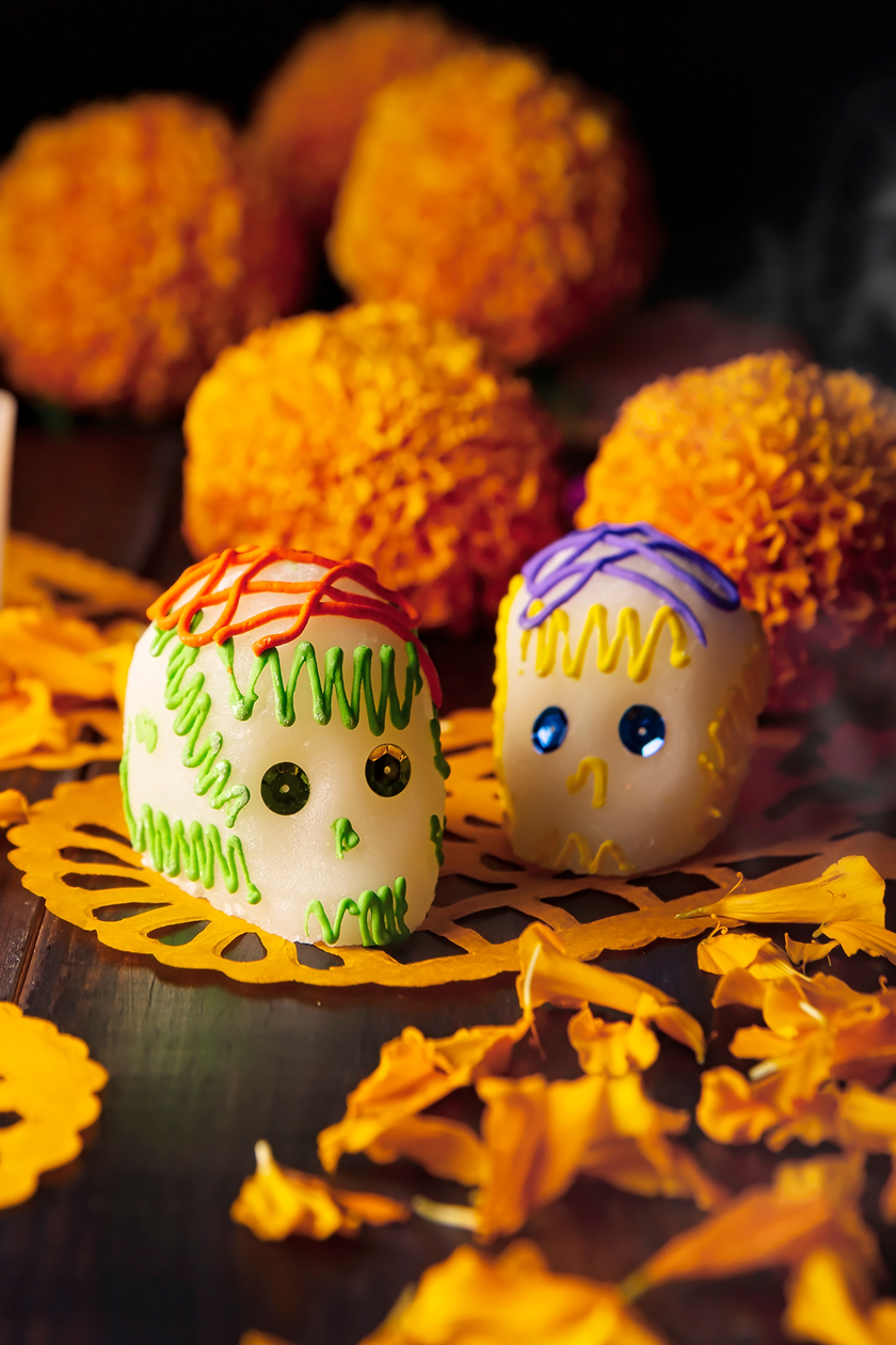 Two decorated sugar skulls on yellow papel picado, surrounded by marigolds.